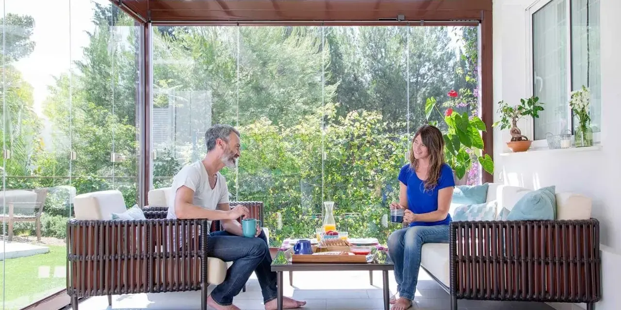 Pareja disfrutando de un café en un **porche acristalado** con vistas a un jardín exuberante.