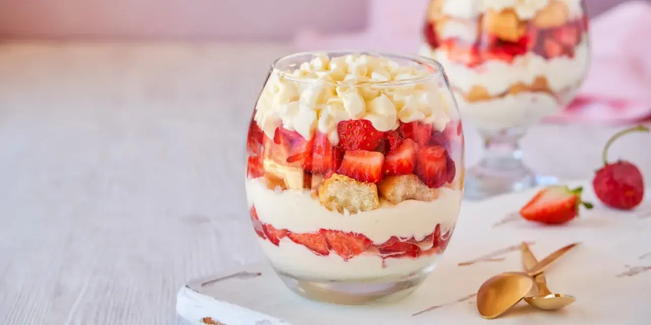 Postre en vaso con capas de crema, fresas y bizcocho, cubierto con trozos de chocolate blanco.