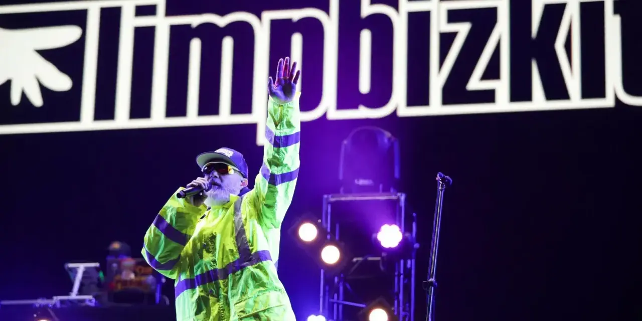Fred Durst z Limp Bizkit na koncercie, w neonowej kurtce, z ręką w górze. Tło to logo zespołu.