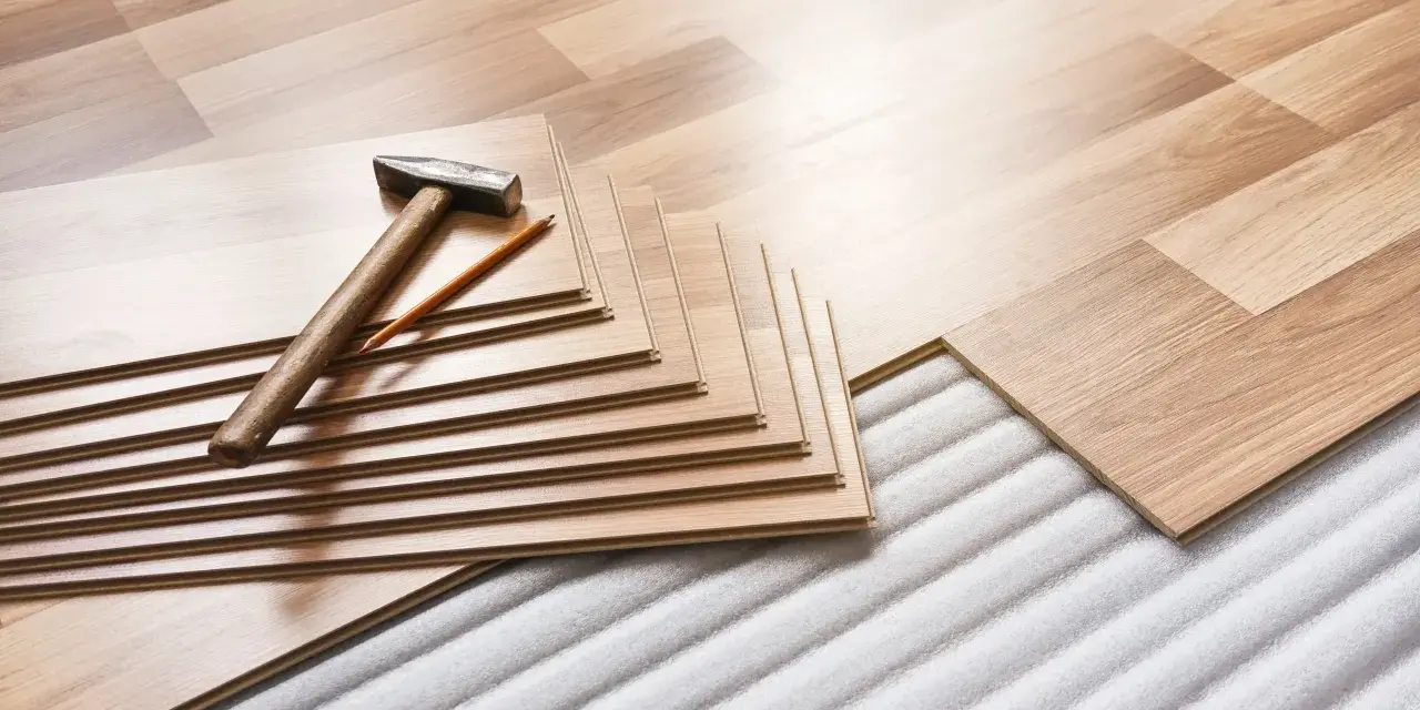 Tablas de suelo laminado apiladas con martillo y lápiz, listas para aislar suelo frío sobre una base acolchada.