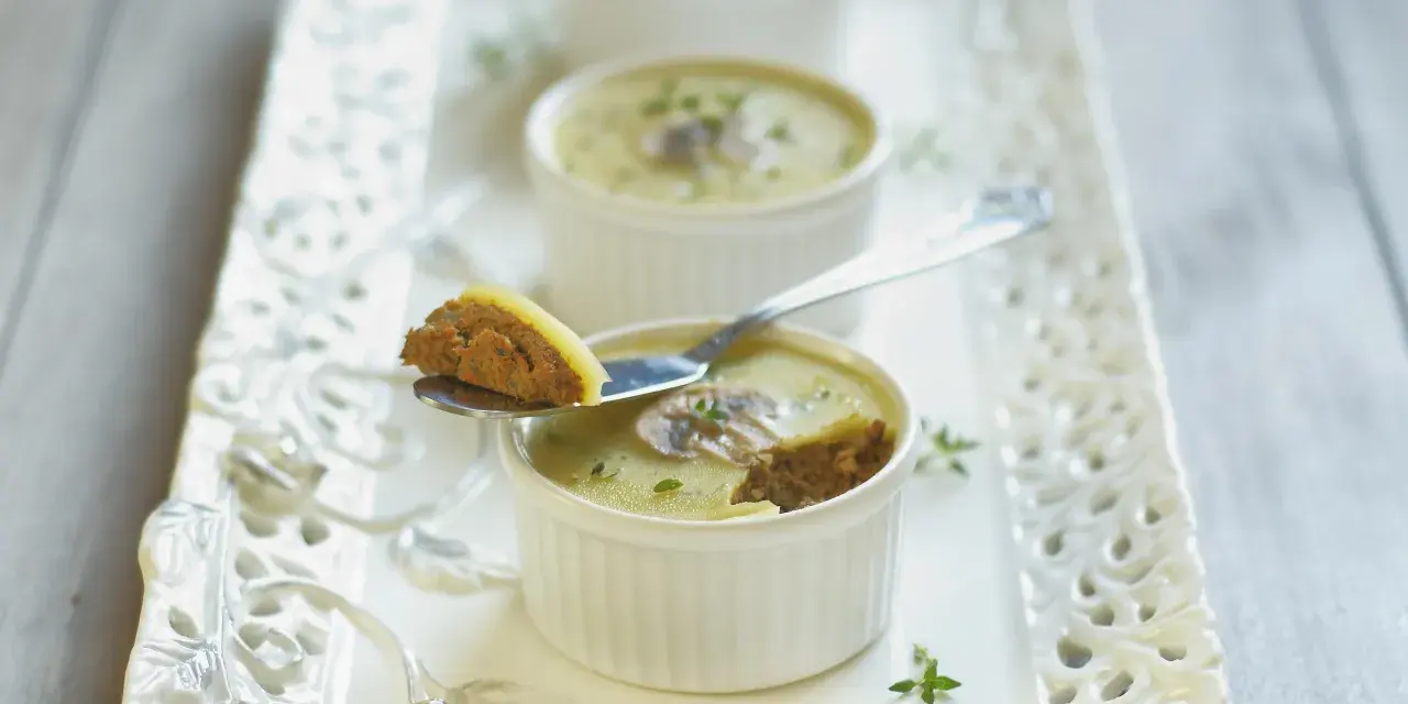 Trois ramequins de foie de volaille, ornés de champignons et de persil, sur un plateau ajouré. Une cuillère présente une bouchée de ce délice.