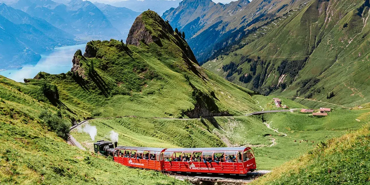 A red Brienz Switzerland train winds through lush green mountains, with a stunning blue lake in the distance.