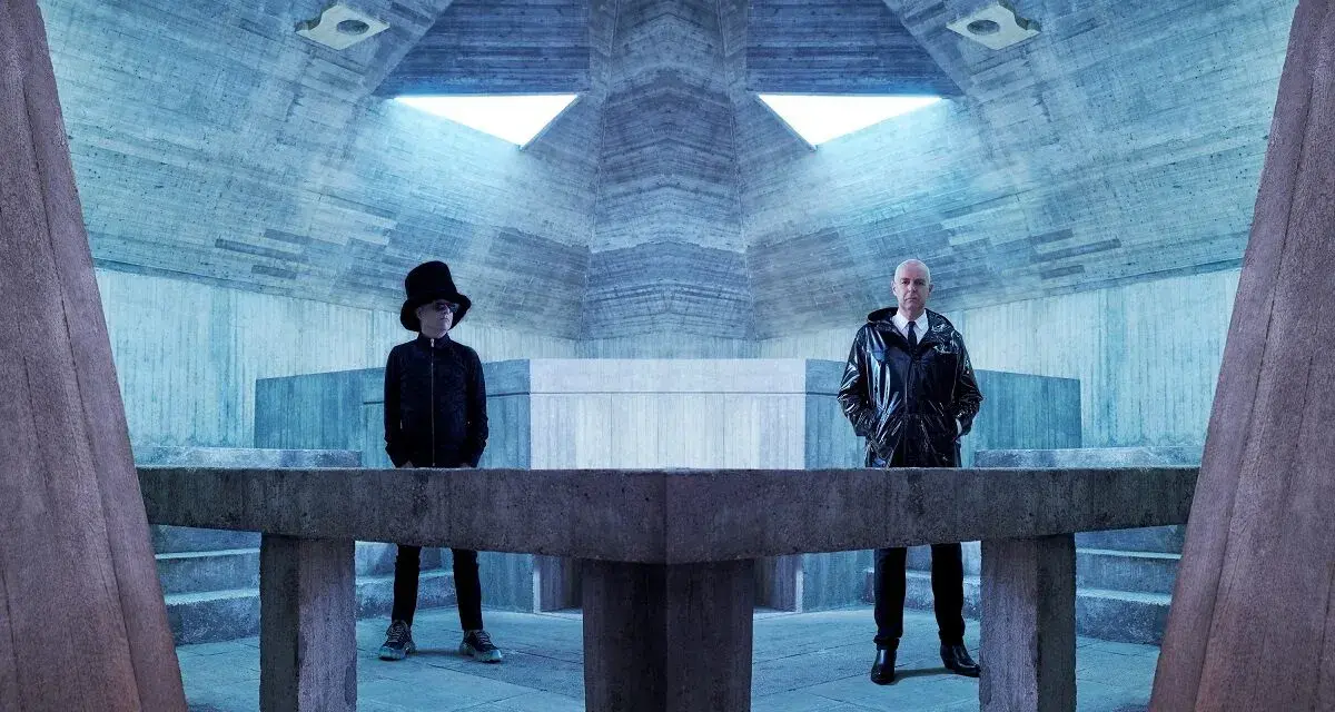 Pet Shop Boys in futuristischer Betonarchitektur. Neil Tennant mit Zylinder, Chris Lowe im glänzenden Mantel.