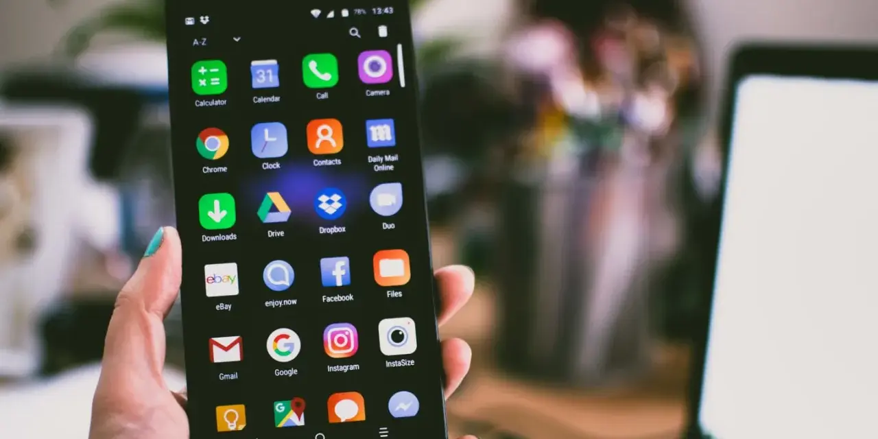Dłoń trzyma smartfon z widocznym ekranem pełnym ikon aplikacji, w tym ikoną wiadomości SMS.