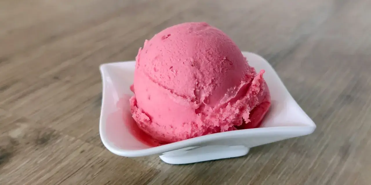 Ein Löffel cremiges Himbeersorbet in einer weißen Schale auf Holzhintergrund.