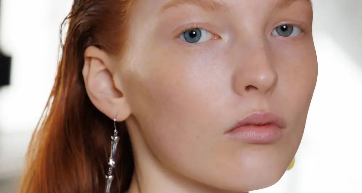 Primo piano di una giovane donna con capelli rossi e occhi azzurri, un neo della bellezza sul viso. Indossa un orecchino pendente.