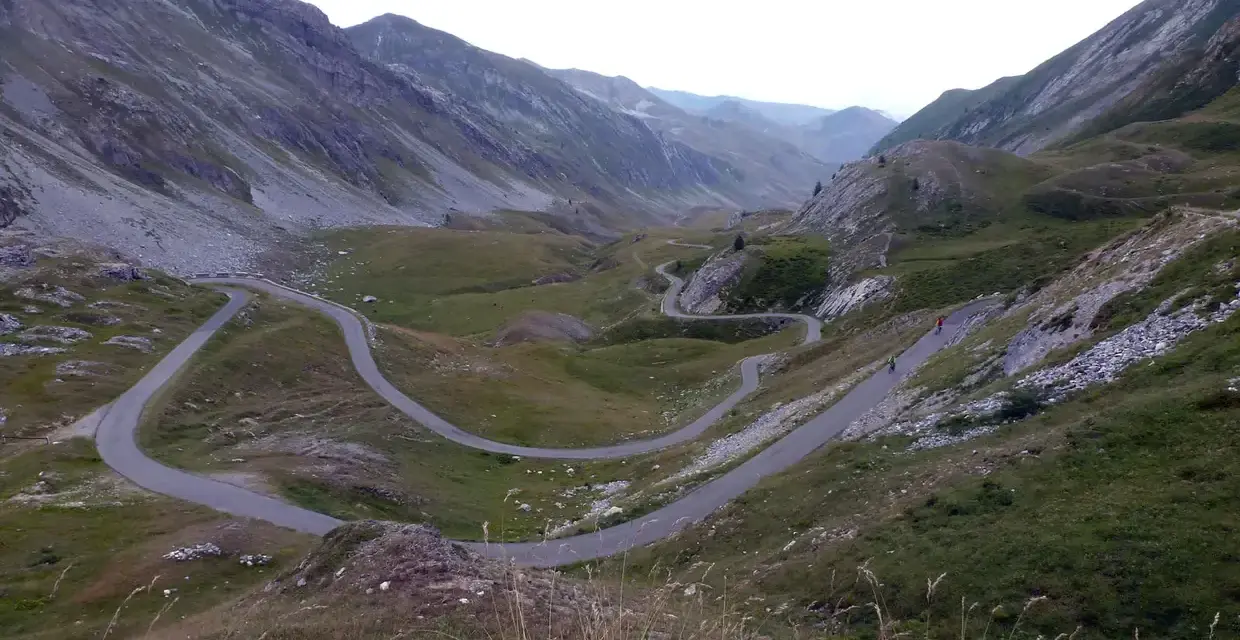 Strada tortuosa che sale verso il colle dei morti, tra montagne verdi e rocciose. Due ciclisti affrontano le curve.