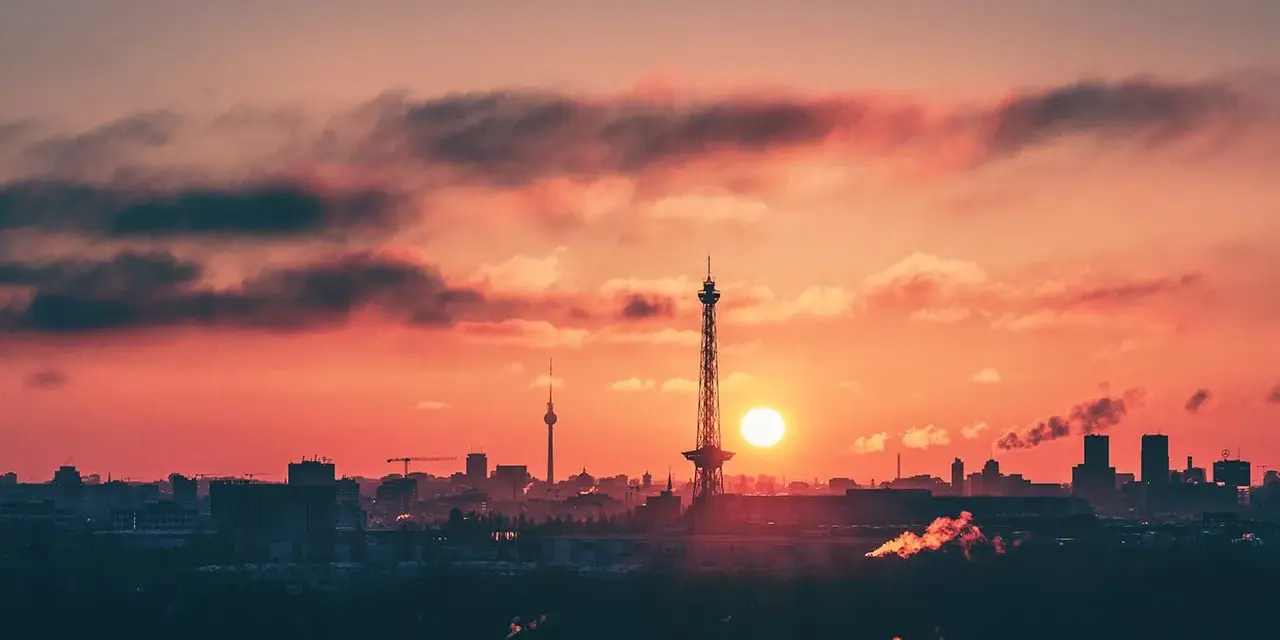 Berlin bei Nacht von oben: Die Sonne geht unter und taucht den Himmel in ein feuriges Rot. Silhouetten von Türmen und Gebäuden zeichnen sich ab.