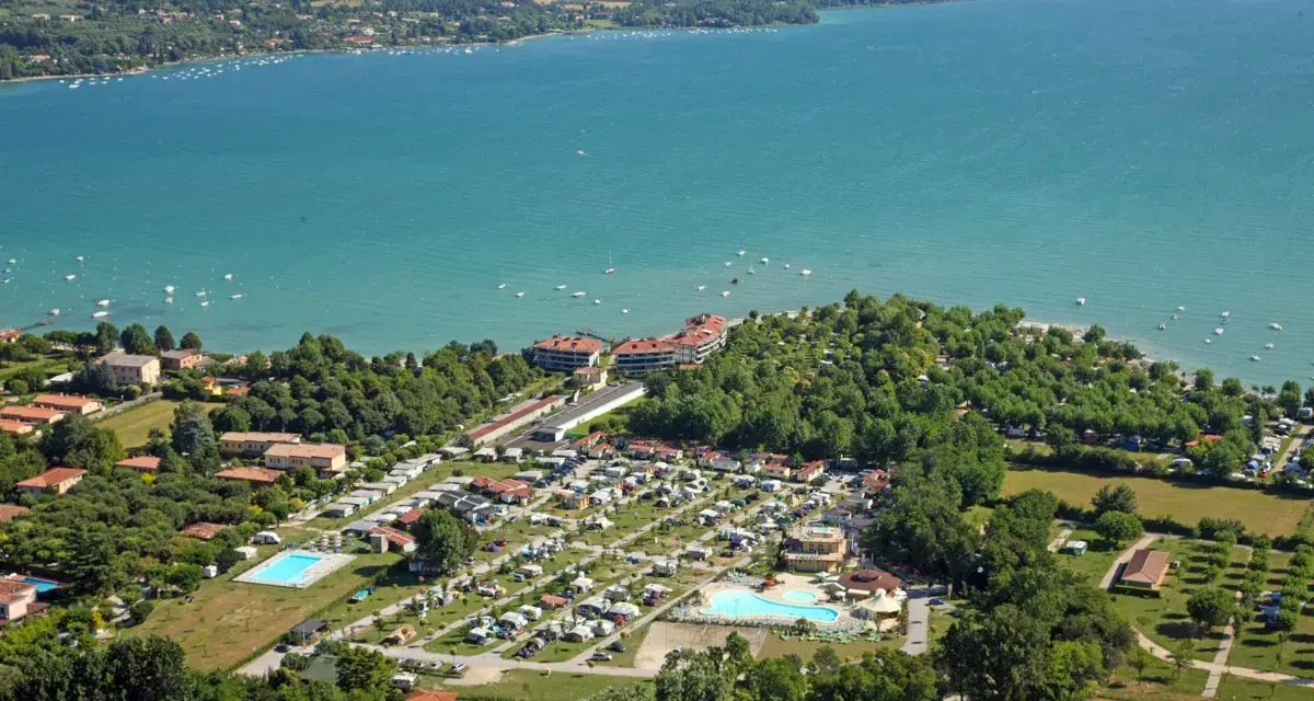 Widok z lotu ptaka na camping Baia Verde nad turkusowym jeziorem, z basenami i licznymi namiotami.