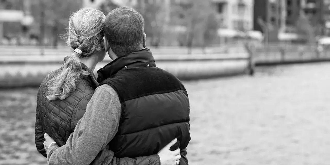 Para przytulająca się nad wodą. Jej ręka obejmuje jego talię, a jego ramiona otulają ją. Delikatny dotyk w relacjach damsko-męskich.
