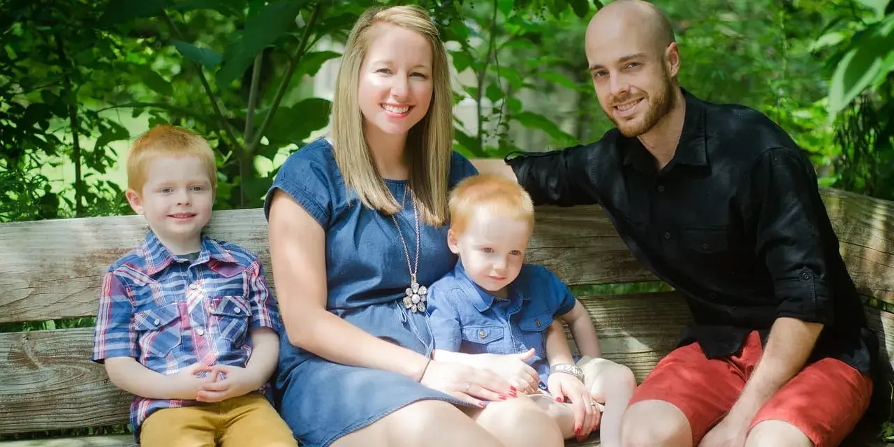 Rodzina na ławce w parku. Dwóch rudowłosych chłopców, mama i tata uśmiechają się. Młodszy syn, jakby w uporczywe naśladowanie innych dzieci, siedzi na kolanach mamy.