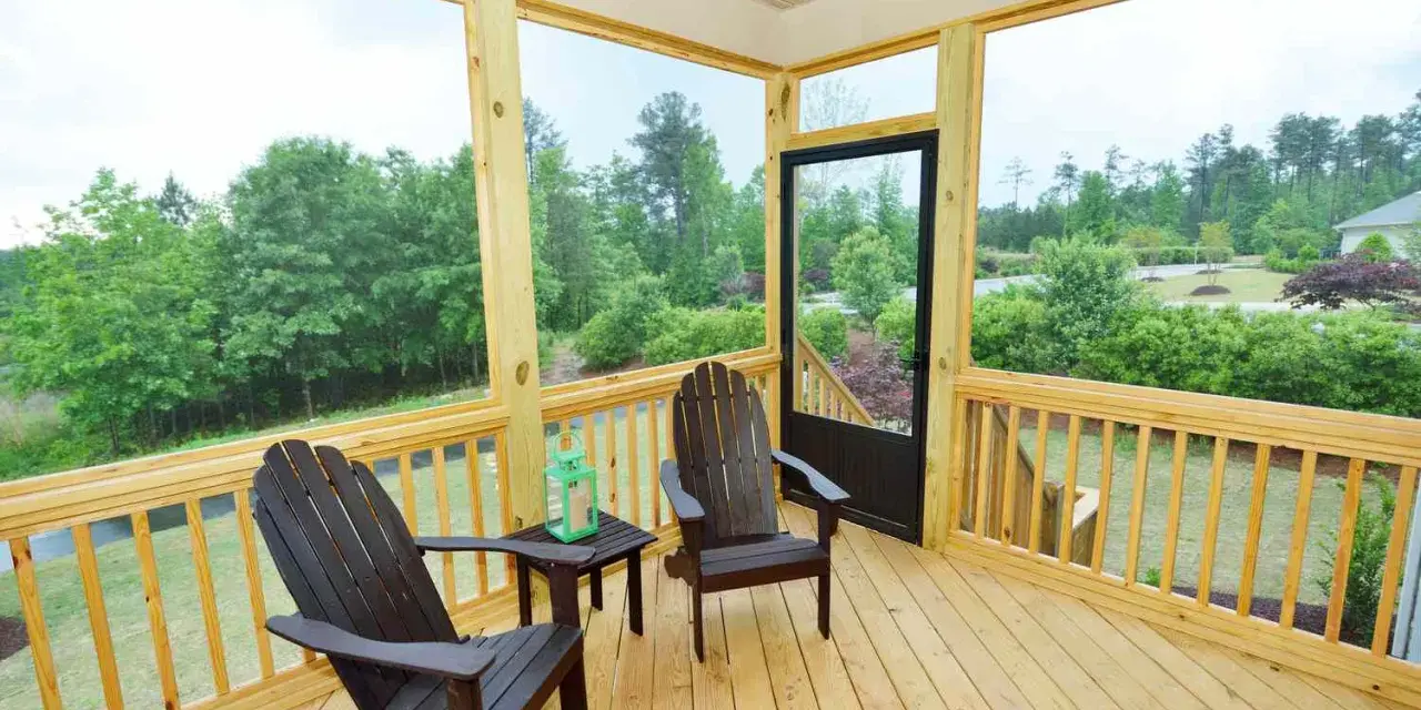 A screened-in porch with two Adirondack chairs, a small table, and a green lantern. The porch offers a view of lush trees and a suburban landscape.