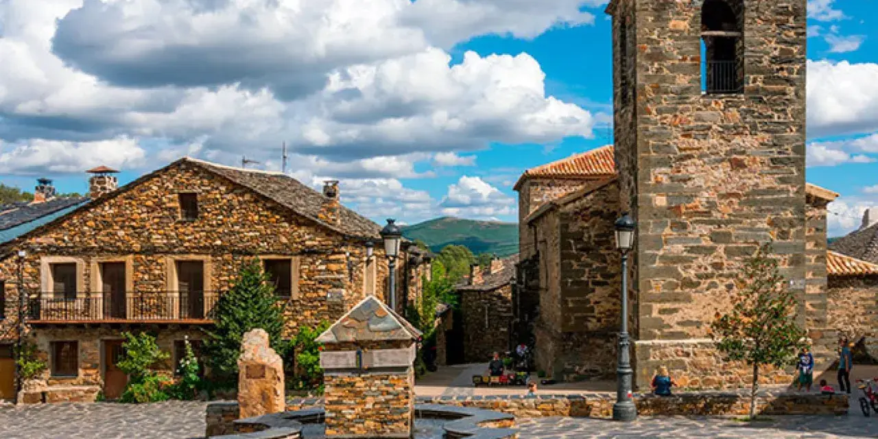 Plaza con fuente de piedra y campanario en un pueblo de la ruta negra Guadalajara. Casas de piedra y cielo azul con nubes.