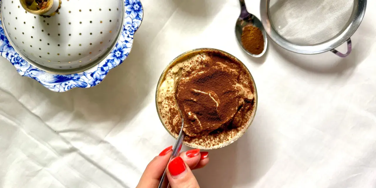 Deliziosa crema al caffè in bicchiere, cosparsa di cacao. Accanto, un elegante contenitore bianco e oro e un cucchiaino con cacao.