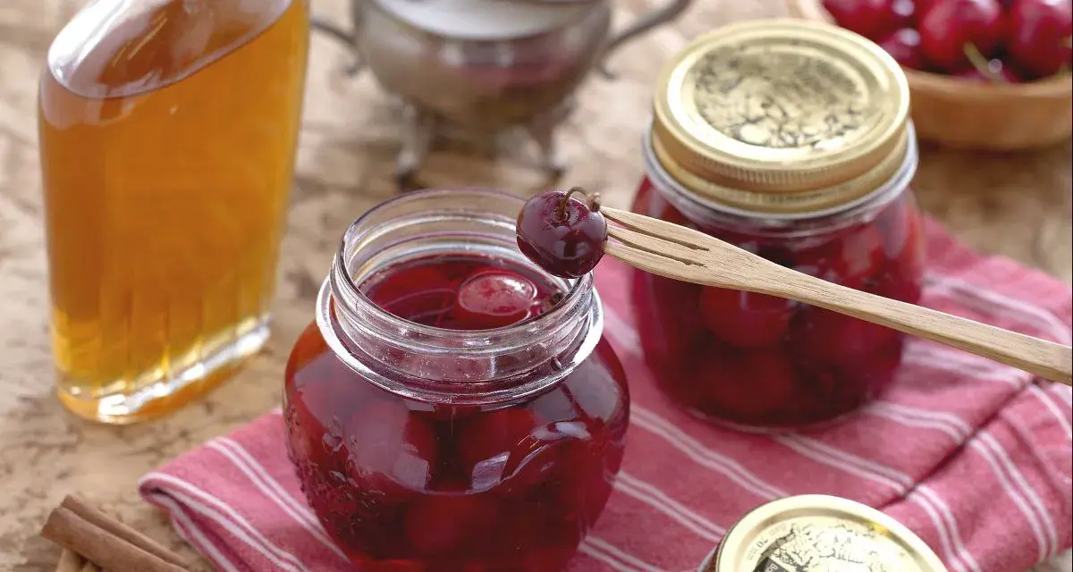 Frascos de mermelada de cerezas casera, una botella de licor y canela en rama. Una cereza en un tenedor de madera lista para probar.