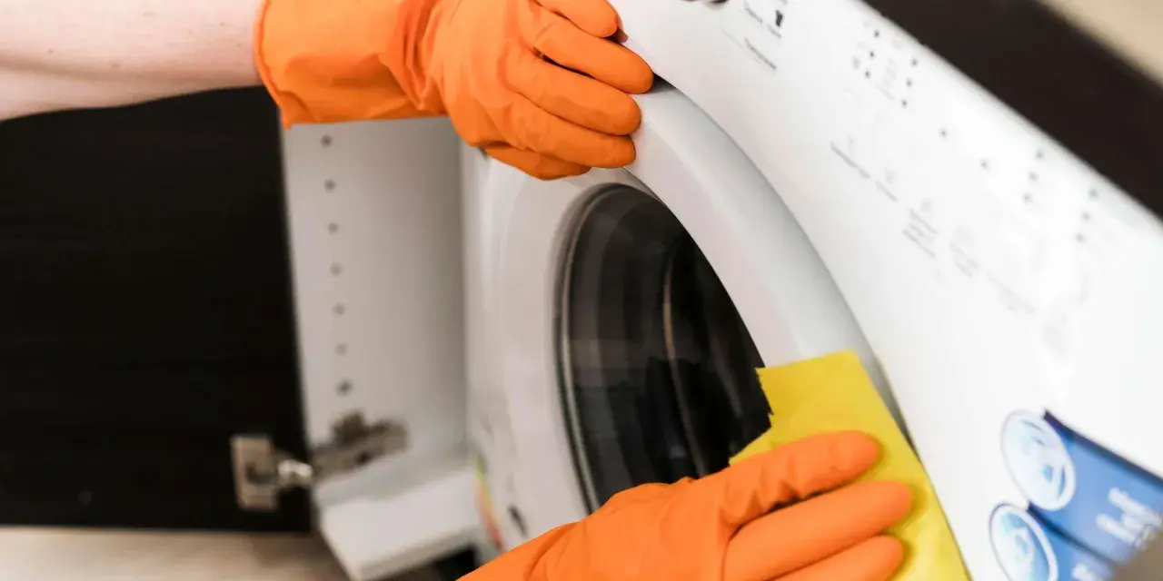 Hände in orangen Handschuhen reinigen die Waschmaschine. Ein gelber Lappen wischt über die Oberfläche, um sie sauber zu halten.