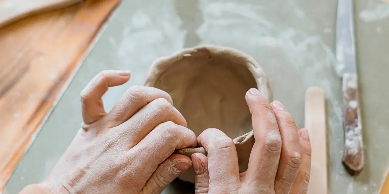 Mani che modellano argilla per creare una ciotola. Utensili da ceramica e un coltello sono visibili. Un ottimo punto di partenza per chi cerca dove trovare l'argilla.