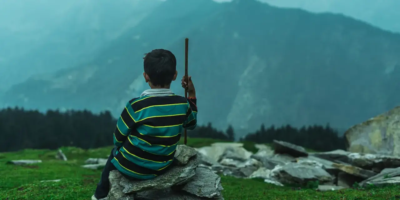 Junge sitzt auf Steinen, blickt in nebelverhangene Berge. Ein Stock in der Hand, wie für einen meditationstext zum vorlesen.