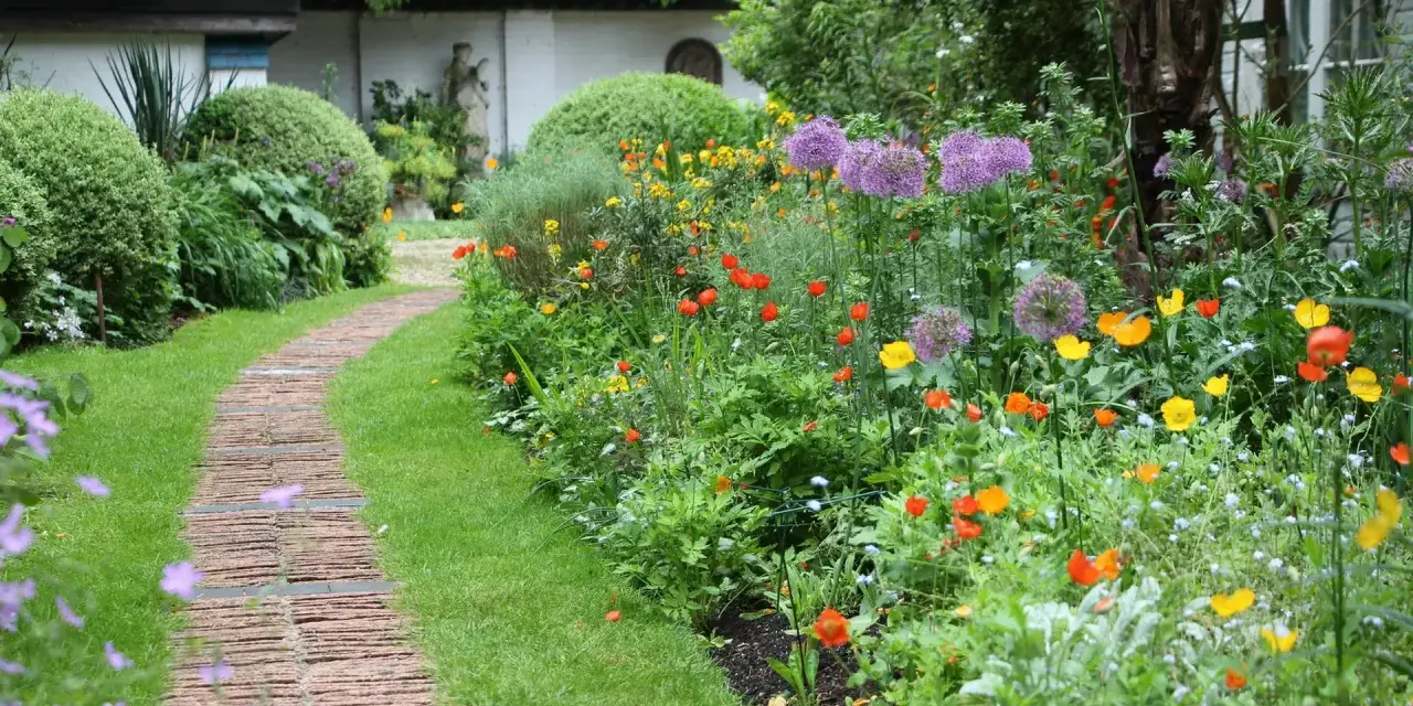 Ein geschwungener Weg aus Ziegelsteinen schlängelt sich durch einen üppigen bauerngarten mit leuchtenden Blumen und grünem Rasen.