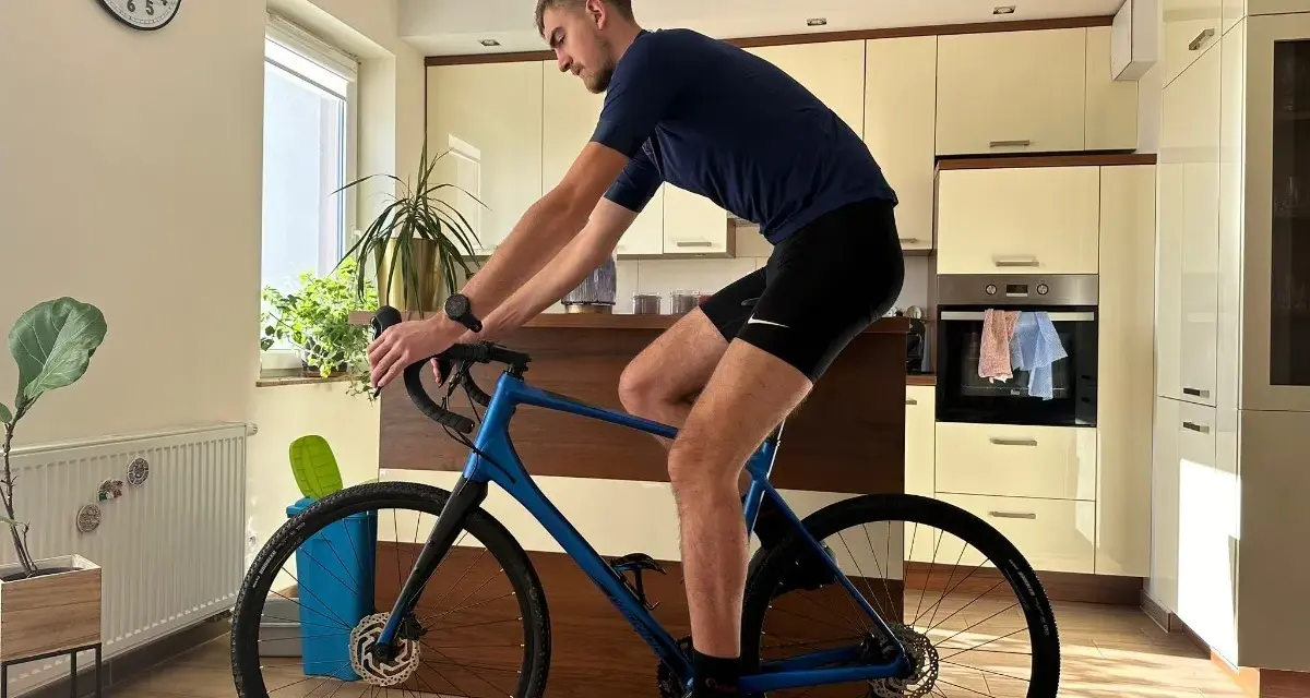 Mężczyzna w pozycji na rowerze gravel, trenuje w domu. W tle kuchnia i zegar.