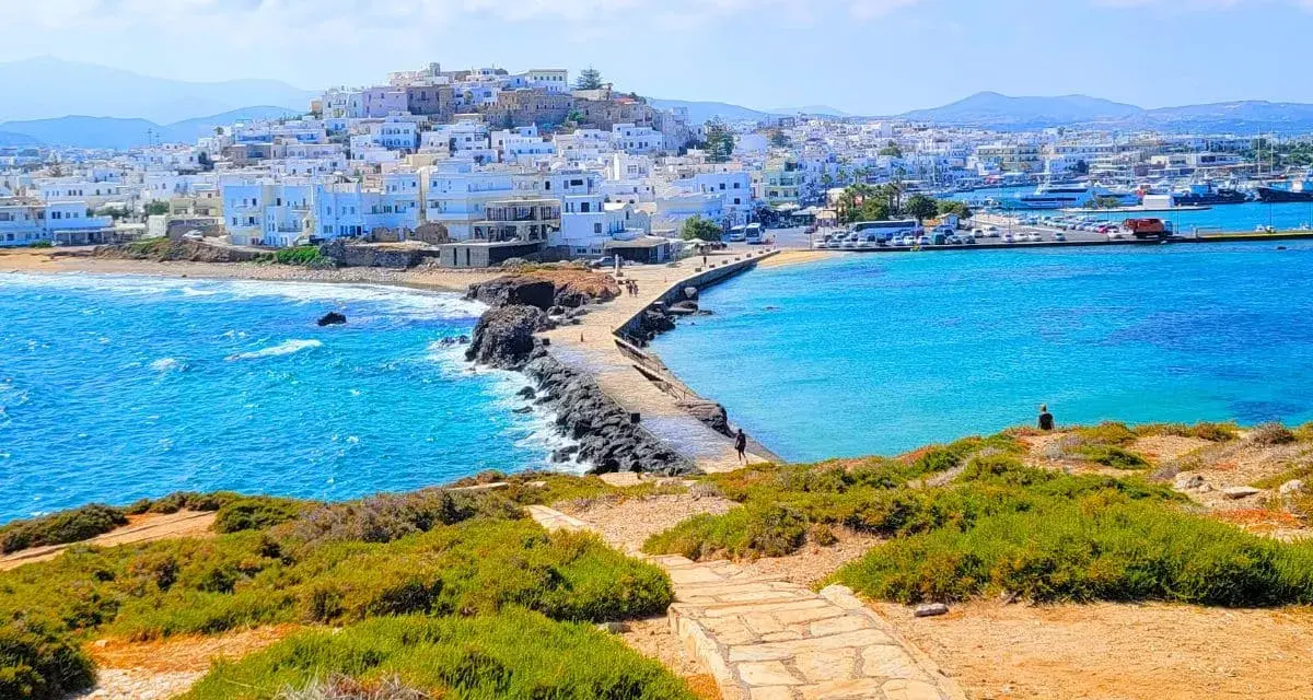 A stone path leads to a causeway connecting to the whitewashed town of Naxos, Greece, with vibrant blue waters lapping at the shore.