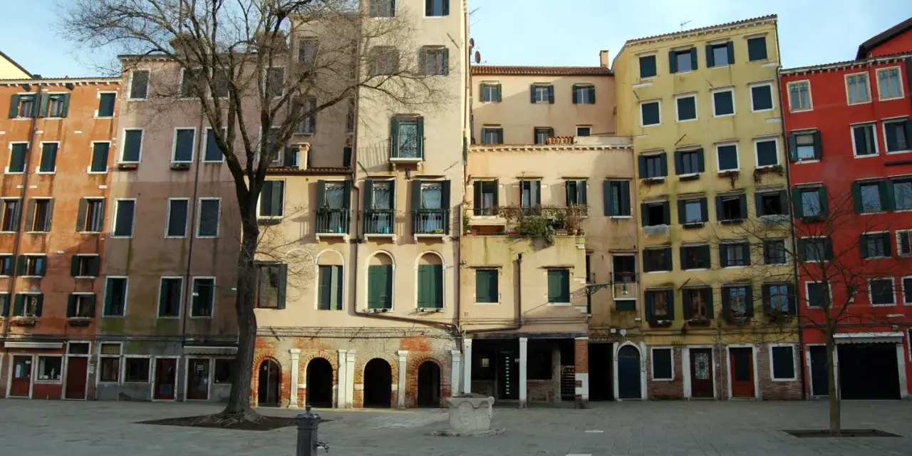 Edifici colorati nel ghetto ebraico di Venezia, con un albero spoglio e una fontana nella piazza lastricata.