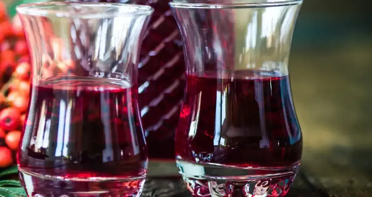 Dwa kieliszki wypełnione rubinową nalewką z czeremchy, obok butelka i kiść owoców.