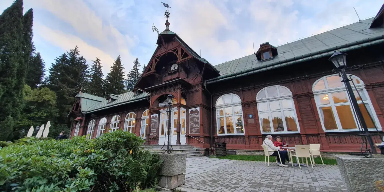 Drewniany pawilon z zegarem i karłowata studzienka przed wejściem. Na zewnątrz siedzą ludzie przy stoliku.