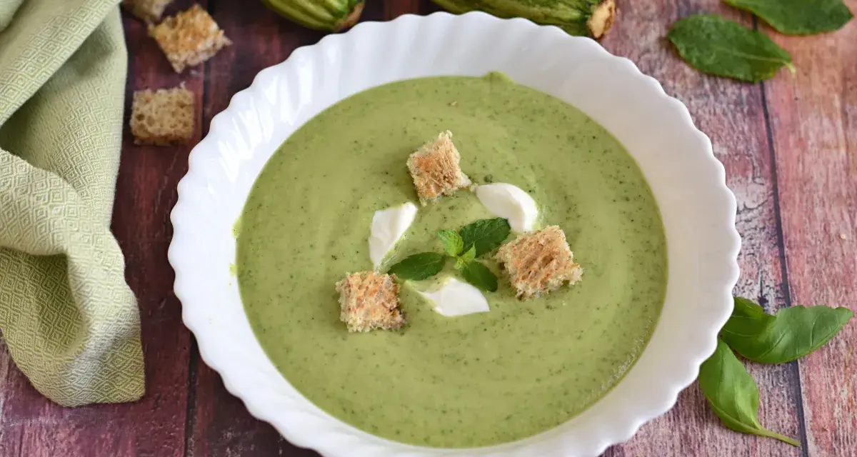 Crema fredda di zucchine servita in una ciotola bianca, guarnita con crostini, panna e menta. Fresca e invitante.