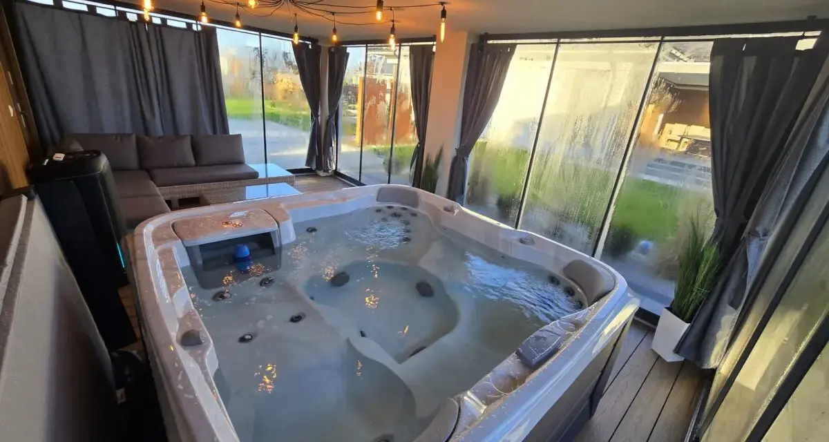 Relaks w jacuzzi pod ciepłym światłem lamp w przeszklonym tarasie zimowym. Obok wygodna sofa i stolik.