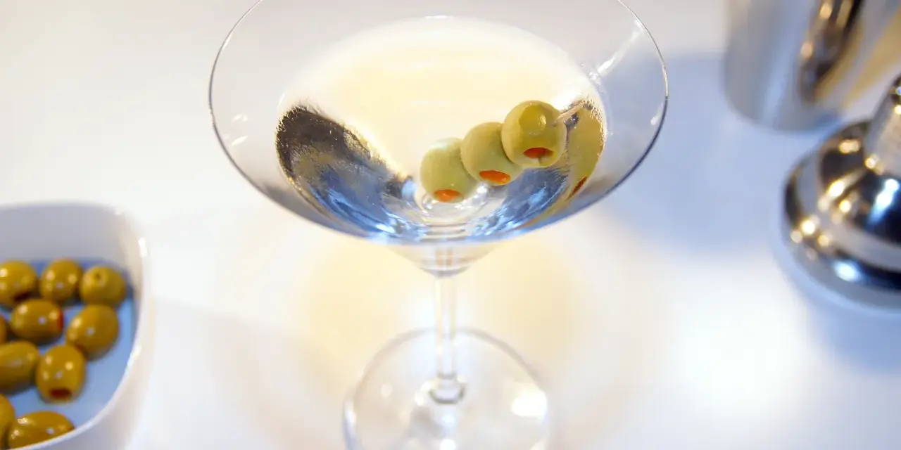 Kto wybierze Dirty Martini czy klasyczne Dry Martini? Kieliszek z oliwkami i drinkiem na białym tle.