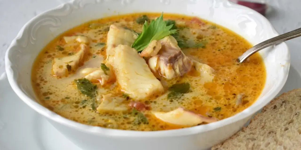 Sopa de pescado casera en un cuenco blanco con trozos de pescado, perejil y una rebanada de pan.