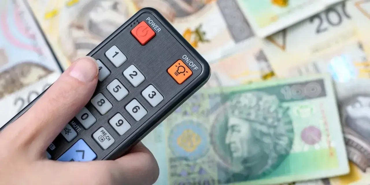 Dłoń trzyma pilota, który steruje kanałem technologicznym, na tle polskich banknotów.
