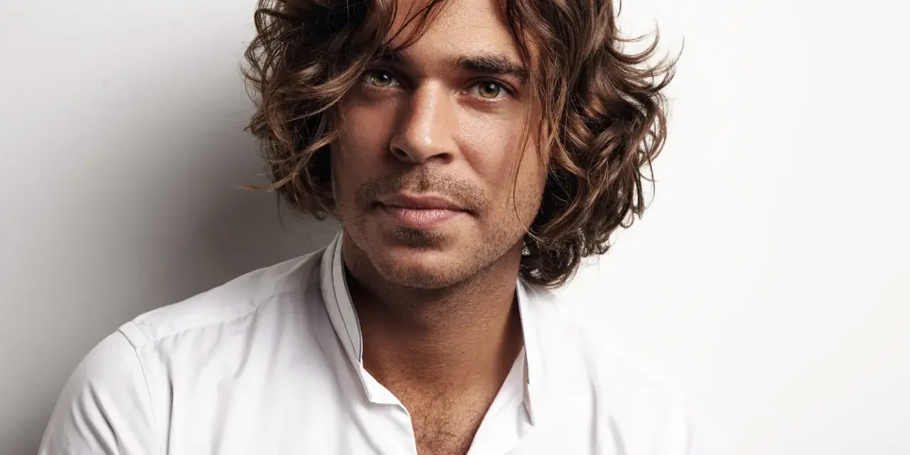 Hombre con cabello castaño ondulado, un ejemplo de cortes de pelo hombre medio largo, viste camisa blanca.