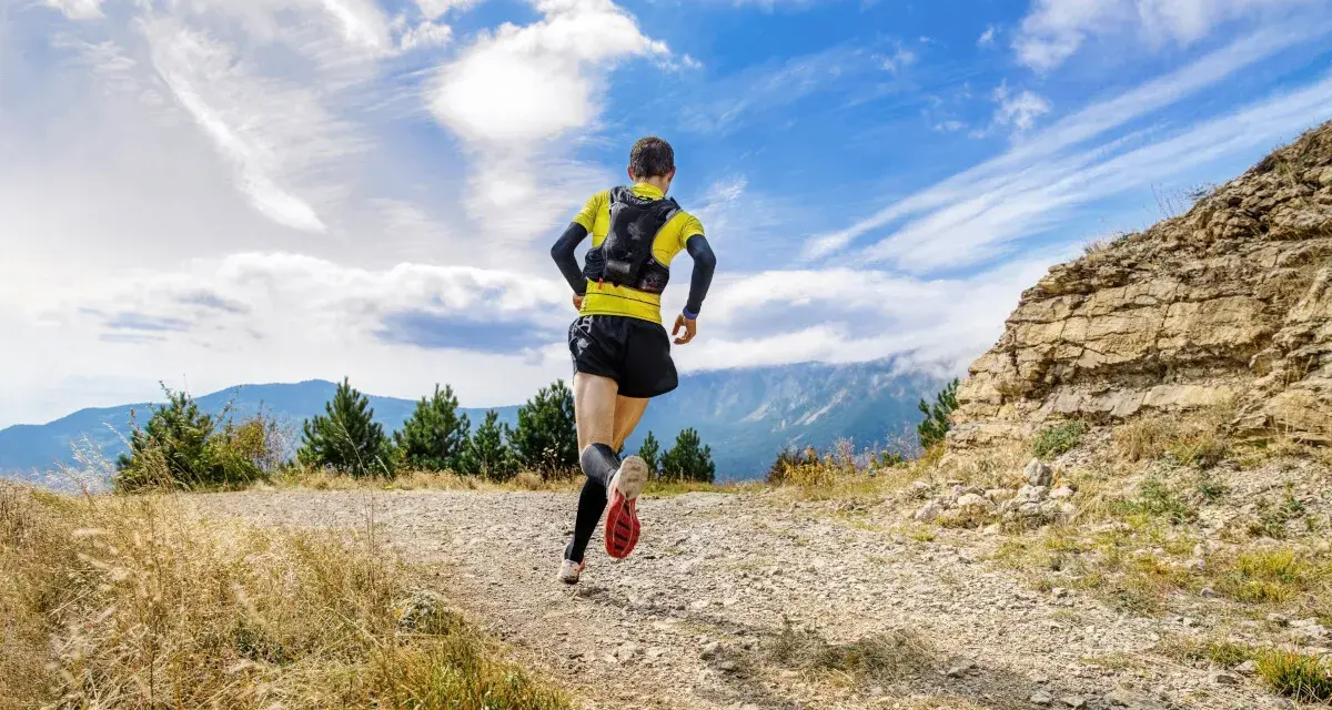 Biegacz w żółtej koszulce pokonuje kamienisty szlak. W tle góry i błękitne niebo z chmurami. To prawdziwe wyzwanie na rykowisko ultra trail.