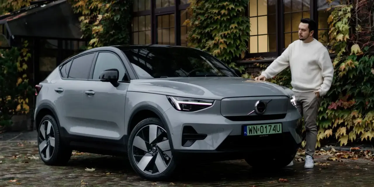 Szary Volvo C40 z czarnym dachem stoi na mokrej, brukowanej nawierzchni. Mężczyzna w białym swetrze opiera się o maskę.
