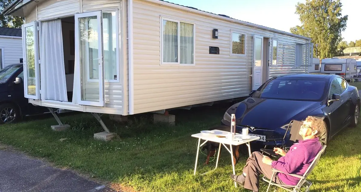 En person njuter av solen vid ett campingbord utanför en husvagn. En Tesla parkerad bredvid. Camping Timrå.