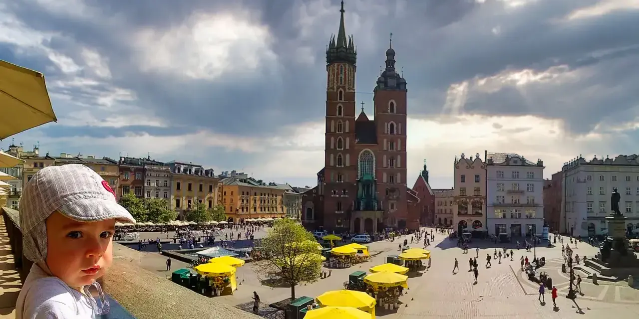 Małe dziecko w czapce patrzy na Rynek Główny w Krakowie, gdzie znajdują się liczne atrakcje dla dzieci w krakowie, w tym kramy z żółtymi parasolami.