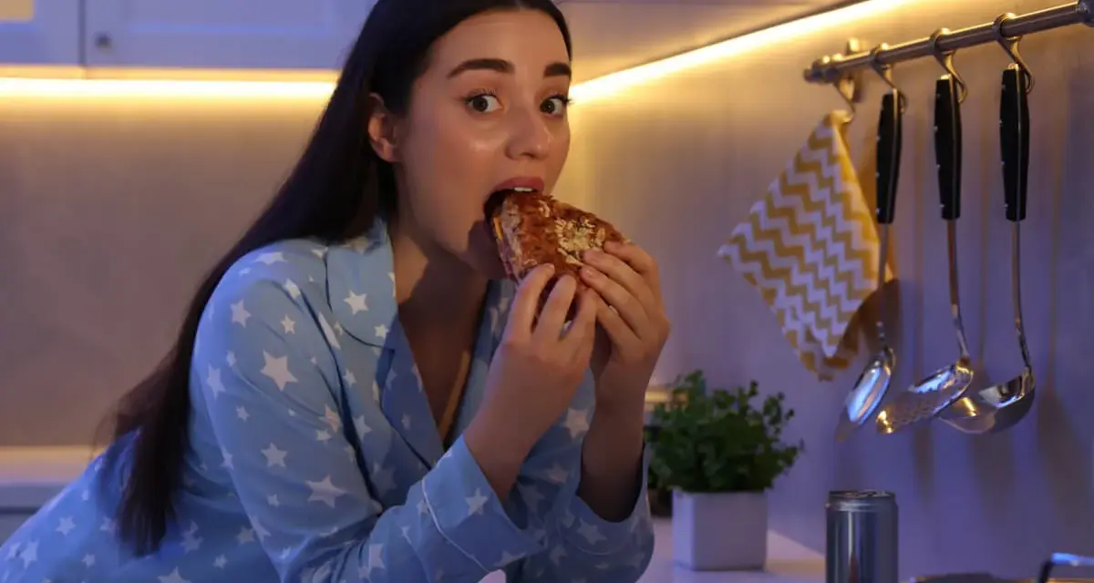 Kobieta w piżamie z gwiazdkami je kanapkę, choć czuje, że jest głodna, ale nie ma apetytu. Na blacie leży też pizza i burger.