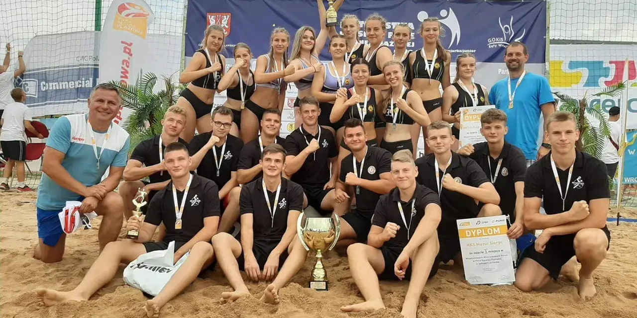 Drużyna MKS Mazur Sierpc świętuje zwycięstwo na piasku, trzymając puchary i dyplomy.