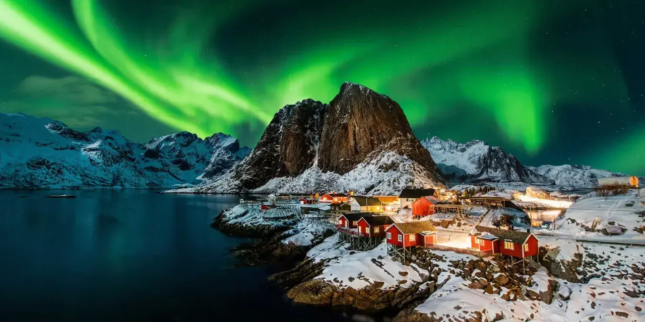 Grön norrsken dansar över snöklädda berg och röda fiskestugor vid en vik i Norge.