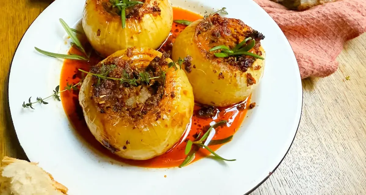 Tres cebollas al horno, rellenas y doradas, nadan en una salsa roja vibrante. Hierbas frescas coronan cada una.