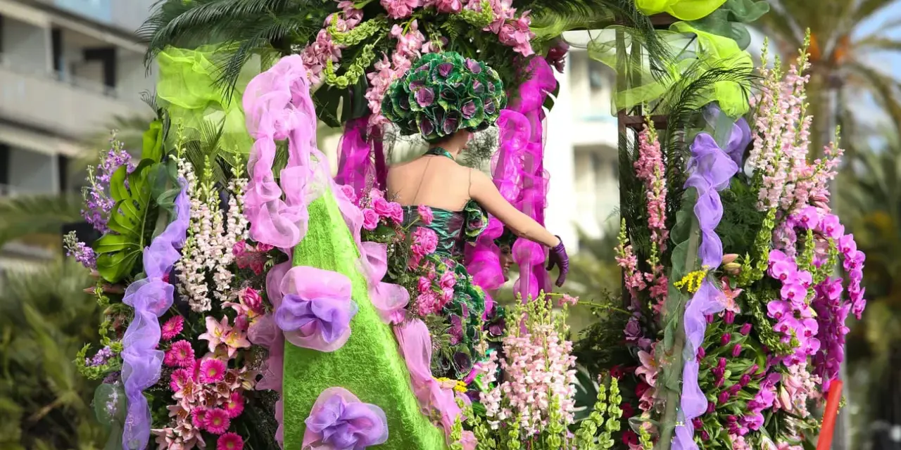 Una figura con un vestido floral y un tocado verde, rodeada de exuberantes flores rosas y moradas, participa en el **carnaval de Nice**.
