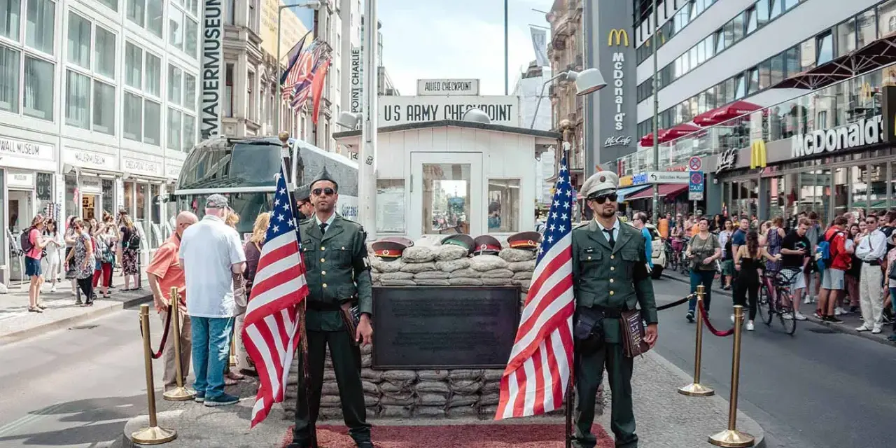 Zwei Männer in Uniformen mit US-Flaggen stehen am Checkpoint Charlie in Berlin. Touristen und ein McDonald's im Hintergrund.