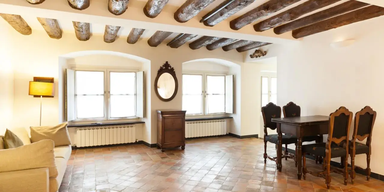 Stary salon z belkami stropowymi i podłogą z terakoty. Kanapa, stół z krzesłami i komoda tworzą przytulną atmosferę.