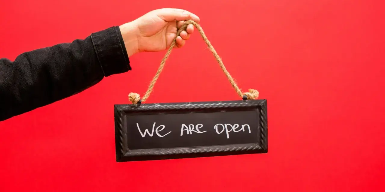 Mano con chaqueta negra sostiene un cartel que dice "We are open", indicando la apertura de un negocio, similar a los tipos de licencia de actividad.