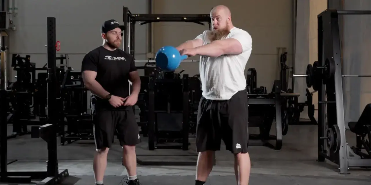Man med skägg utför kettlebell swing varje dag i gym. Bredvid står en tränare.
