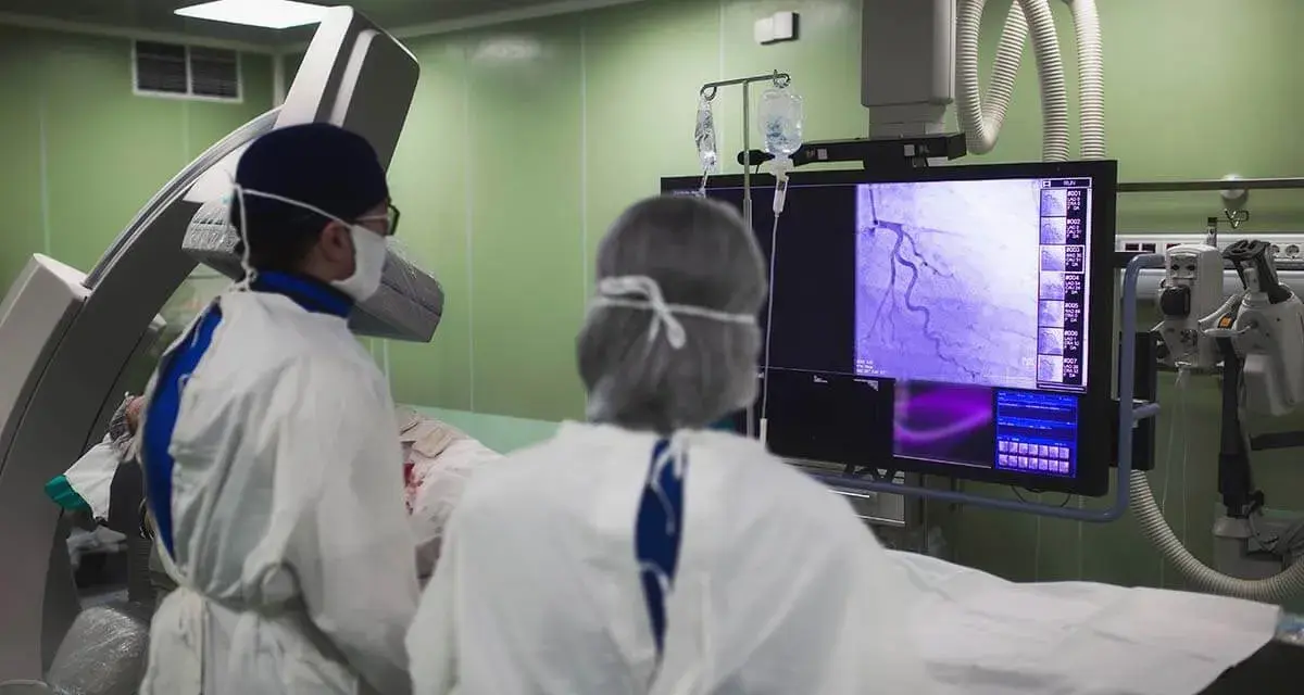 Lekarze w sterylnym stroju analizują obraz z koronarografii na monitorze w sali operacyjnej.