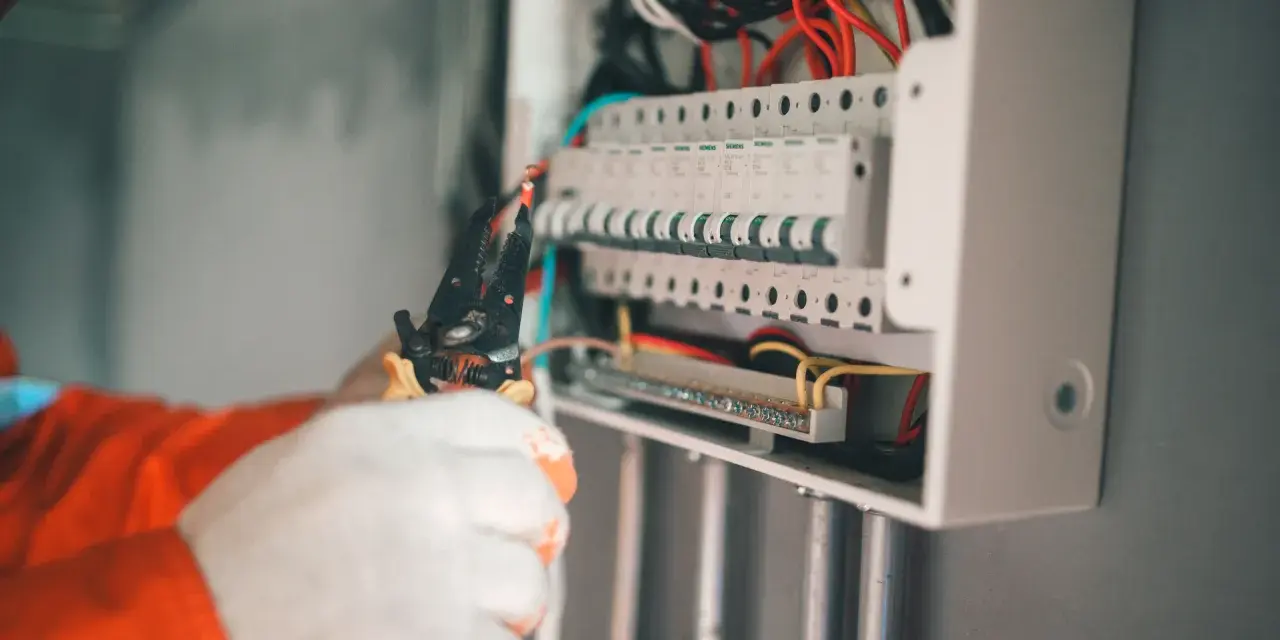 Elektryk w białych rękawicach wywala bezpiecznik w skrzynce, używając szczypiec.