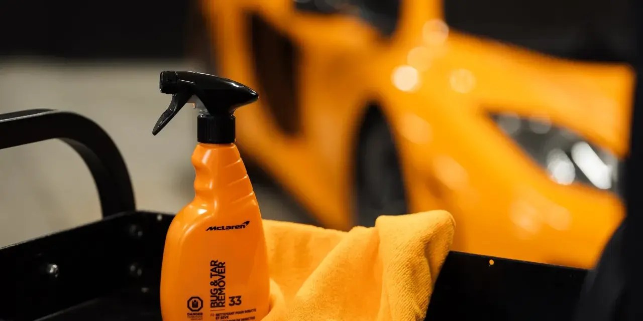 En orange sprayflaska för att ta bort limrester och insekter, bredvid en orange mikrofiberduk. En sportbil i bakgrunden.